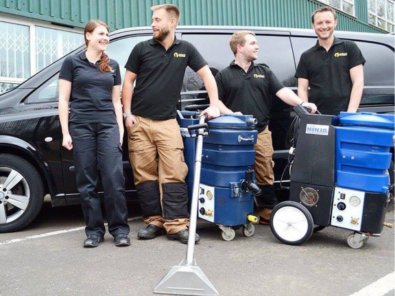 This is a photo of carpet cleaners (three men and one woman) standing in fromt of their black van, with two steam cleaning carpet machines next to them